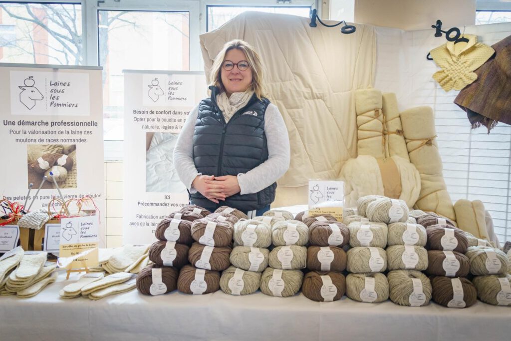Femme souriante devant un stand de "Laines sous les Pommiers", présentant des produits en laine naturelle et durable.
