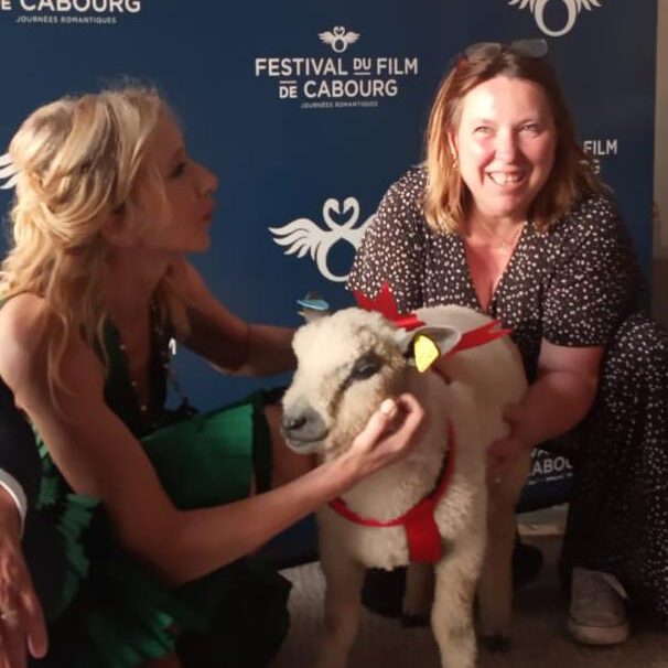 Deux femmes entourent un mouton portant un collier rouge, devant un fond du Festival du Film de Cabourg.
