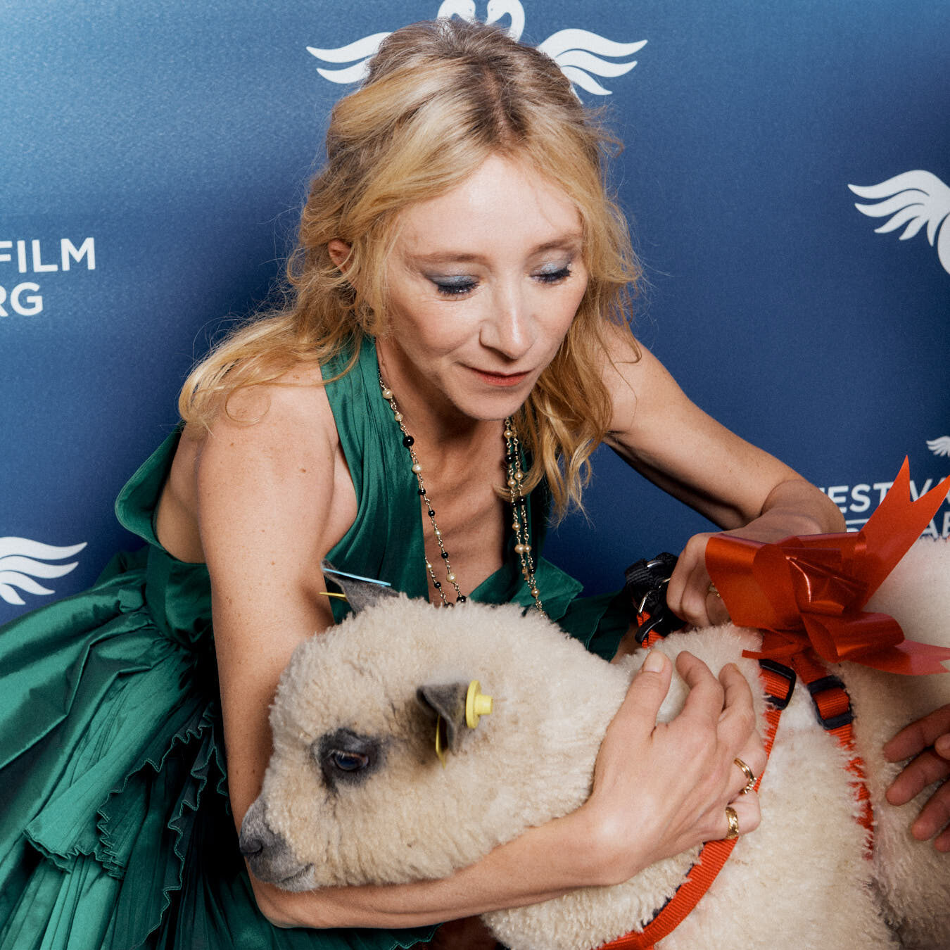 Femme en robe verte tenant un agneau avec un nœud rouge, sur un fond de festival de cinéma.