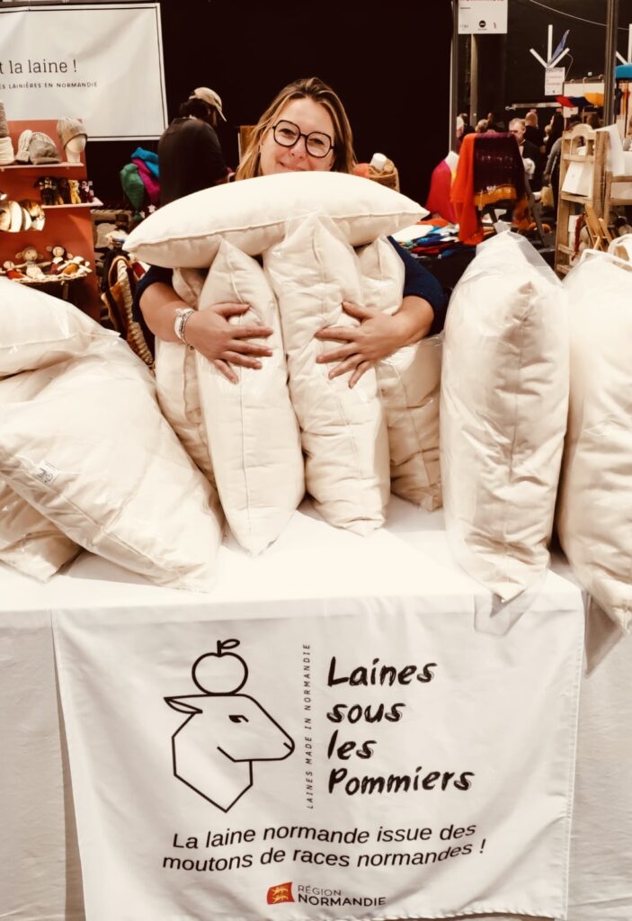 Femme souriante tenant des oreillers en laine, devant un stand de Laines sous les Pommiers, mettant en avant la laine normande.