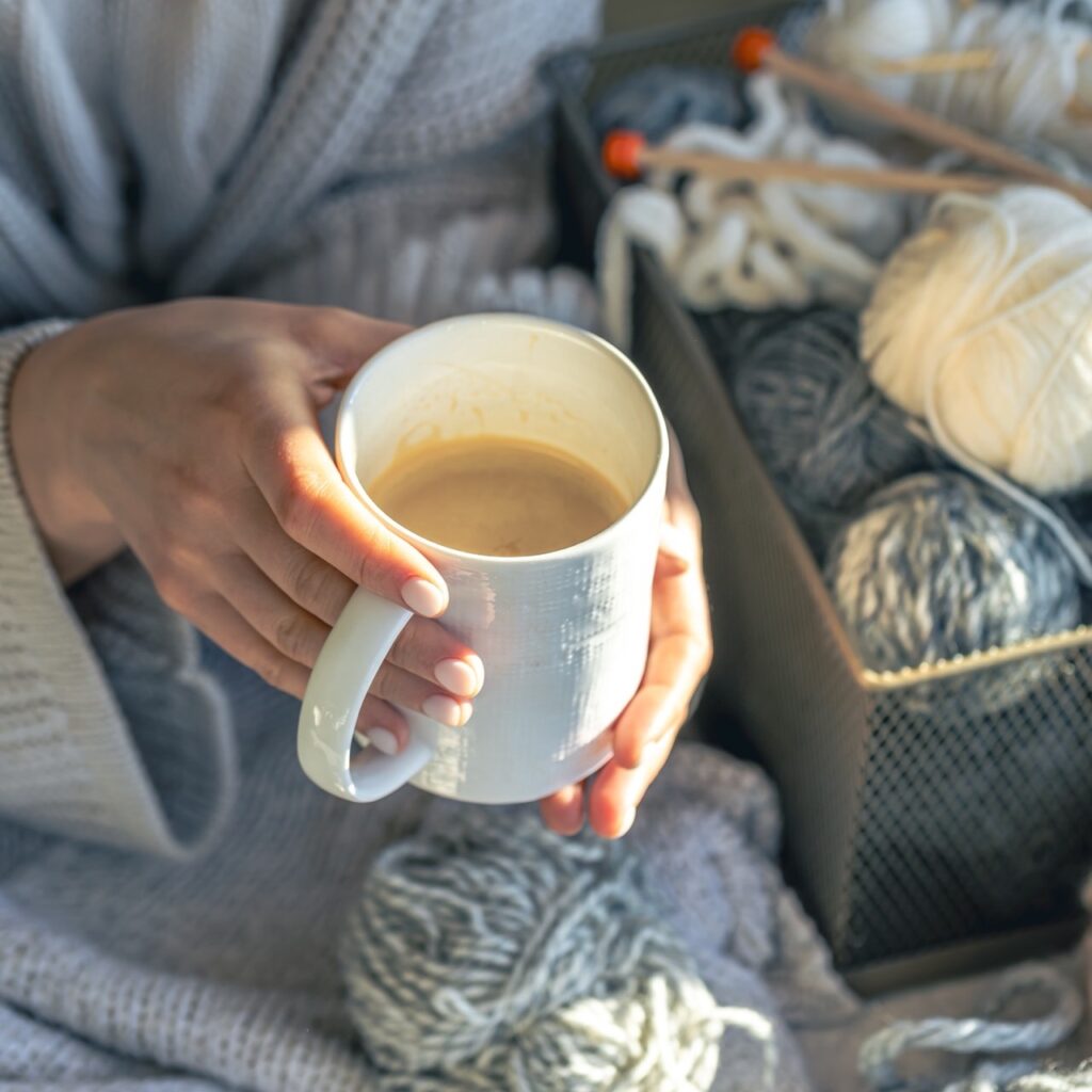 Personne tenant une tasse de café, entourée de pelotes de laine dans un environnement cozy.