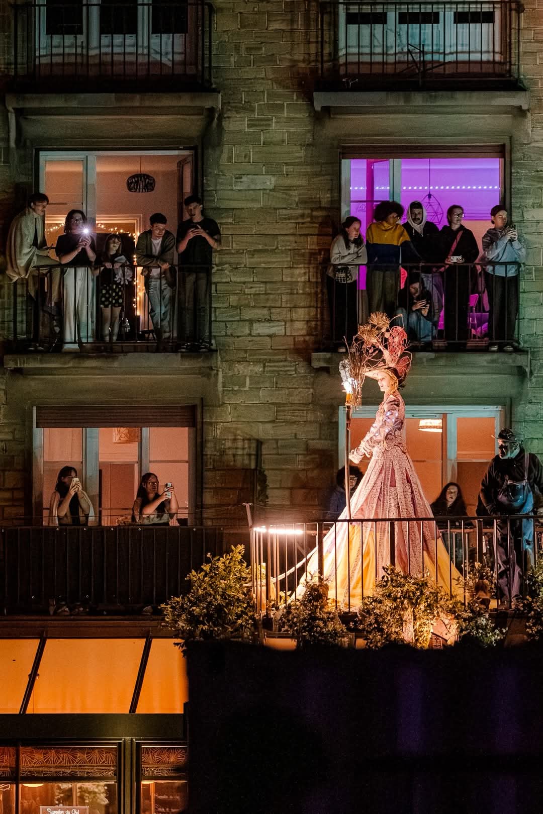 Une grande figure en robe colorée défile sur un balcon, observée par un public enthousiaste dans des appartements voisins.