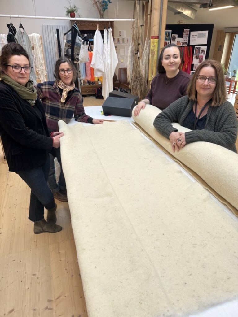 Quatre femmes souriantes présentent un rouleau de laine dans un atelier, entourées de vêtements et de matériaux artisanaux.