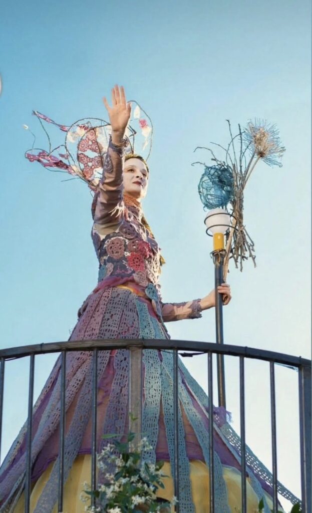 Femme en robe colorée, levant la main, debout sur une plateforme avec un ciel bleu en arrière-plan.