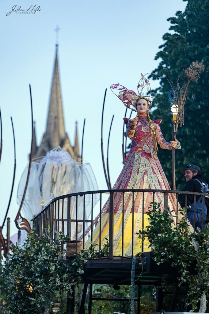 Une femme en costume coloré se tient sur une scène décorée, avec un église en arrière-plan, lors d'un événement festif.