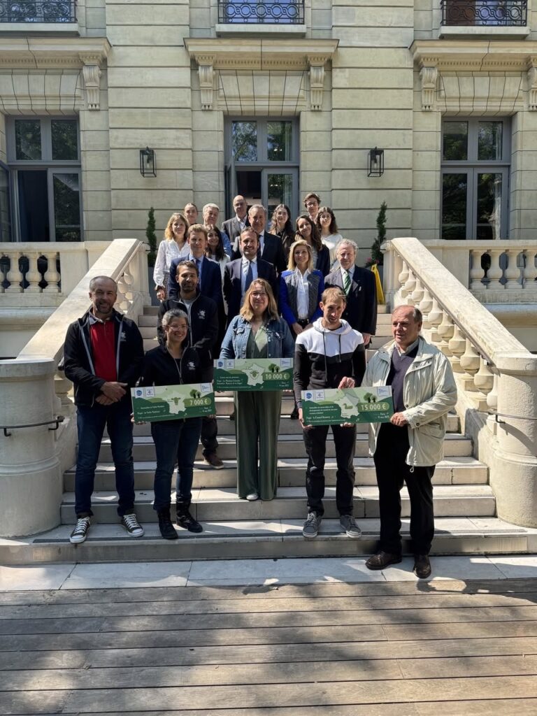 Groupe de personnes posant avec des chèques dans un cadre extérieur architectural, promouvant des initiatives locales.
