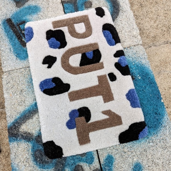 Tapis en laine, motif abstrait avec les lettres "PUT1", sur fond clair, posé sur un sol de béton.