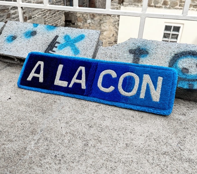 Écusson bleu et blanc avec l'inscription humoristique "A LA CON", posé sur un mur en pierre.