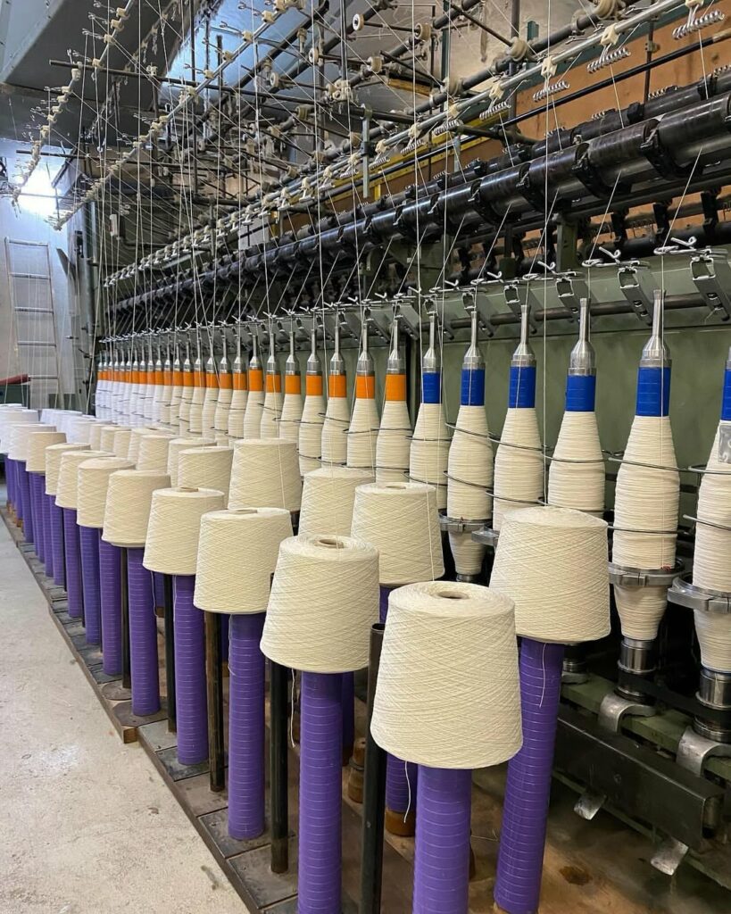 Atelier de production de laine, avec des bobines de fil sur présentoirs et des machines en arrière-plan. Artisanat normand.
