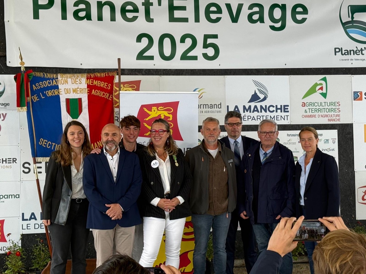 Groupe de personnes posant devant un panneau de l'événement "Planet'Élevage 2025", représentant l'élevage normand et l'artisanat local.